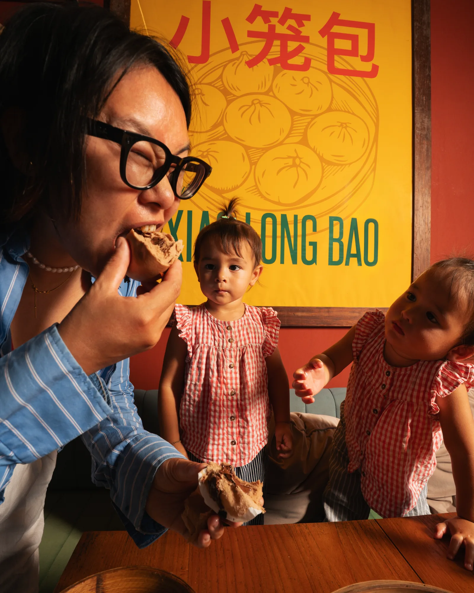 Guest eating a steamed bun beside two children, in front of a Xiaolongbao poster