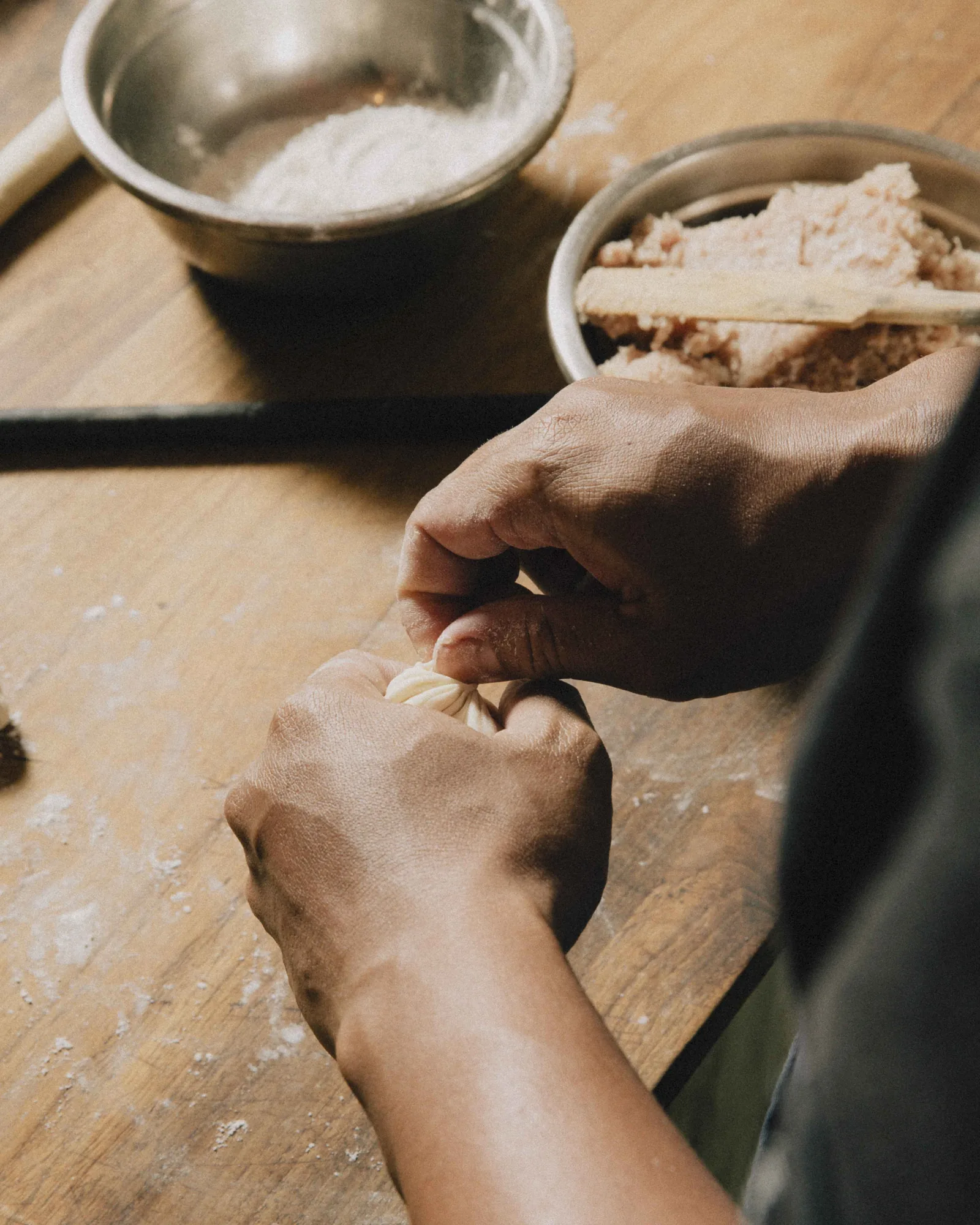 Hands shaping a piece of dough on a flour-dusted wooden prep bench, beside bowls of flour and filling