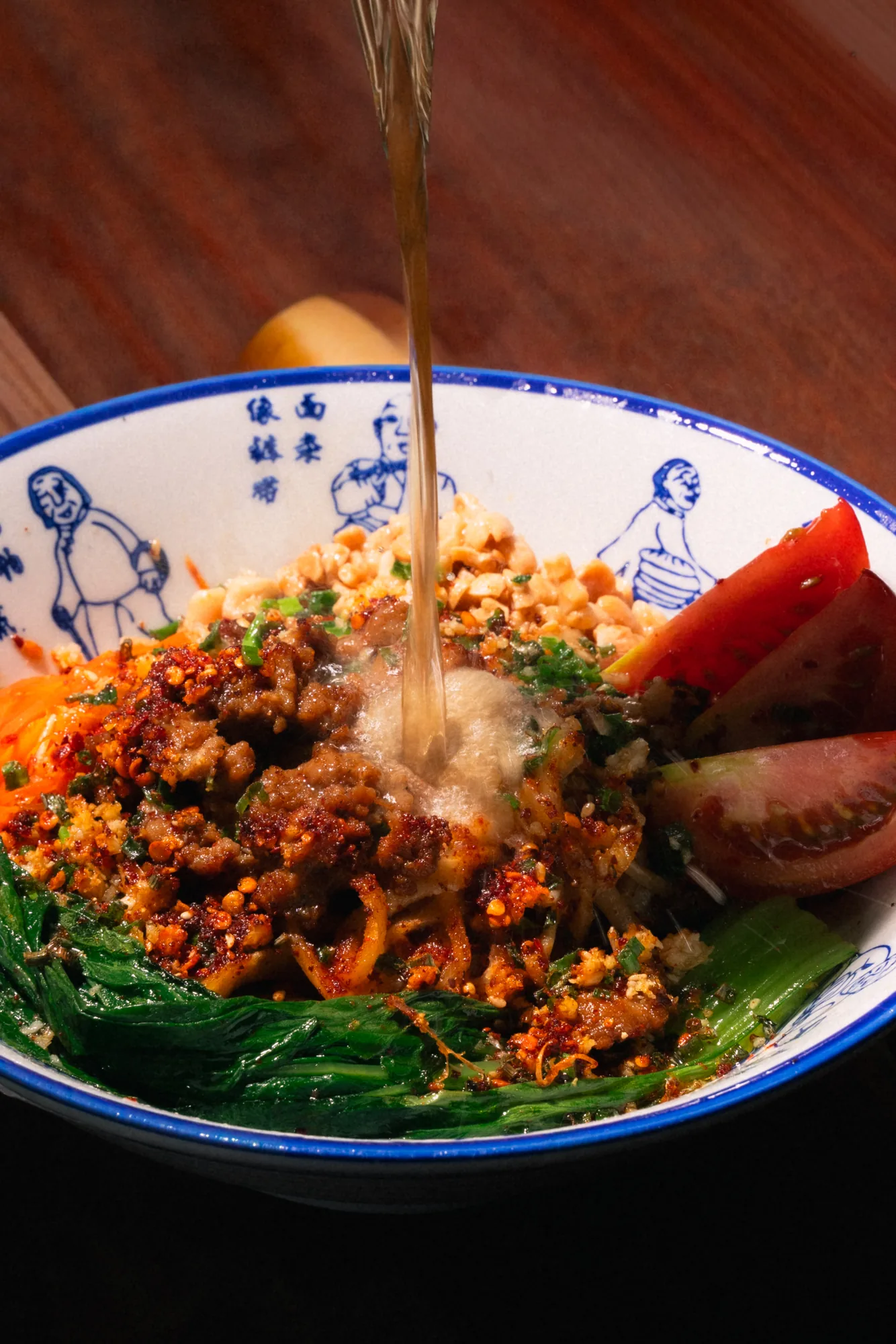 Chilli broth being poured over a bowl of noodles with minced meat and greens